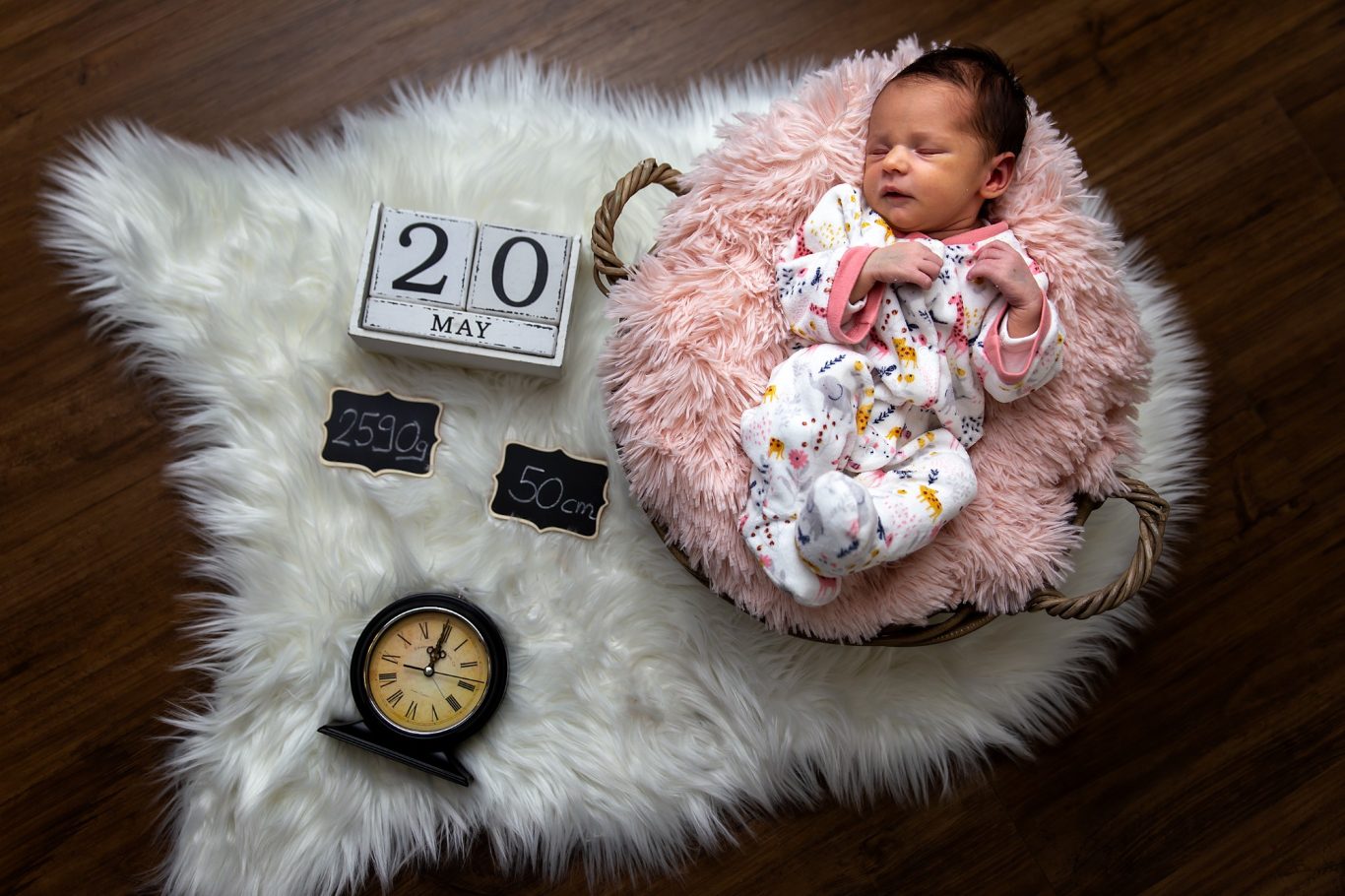 Neugeborenes Baby in einem Korb auf einem weichen Teppich, umgeben von einer Uhr und Datenblock.