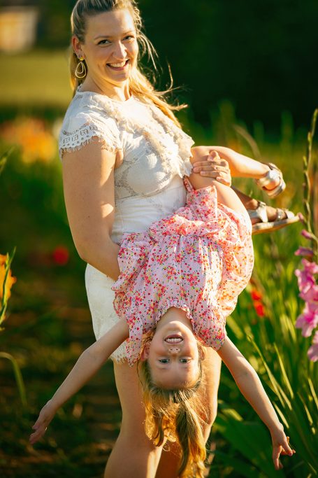 Eine lächelnde Frau hält ein fröhliches Mädchen, das kopfüber hängt, umgeben von Blumen.