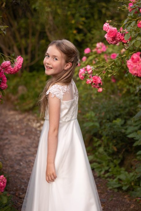 Ein Mädchen in einem weißen Kleid steht im Garten zwischen rosa Rosen.