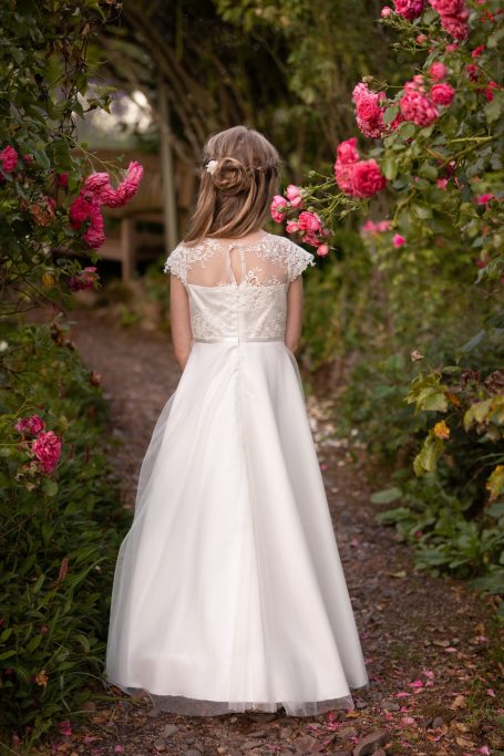Mädchen in einem weißen Kleid steht mit dem Rücken vor blühenden Rosen.