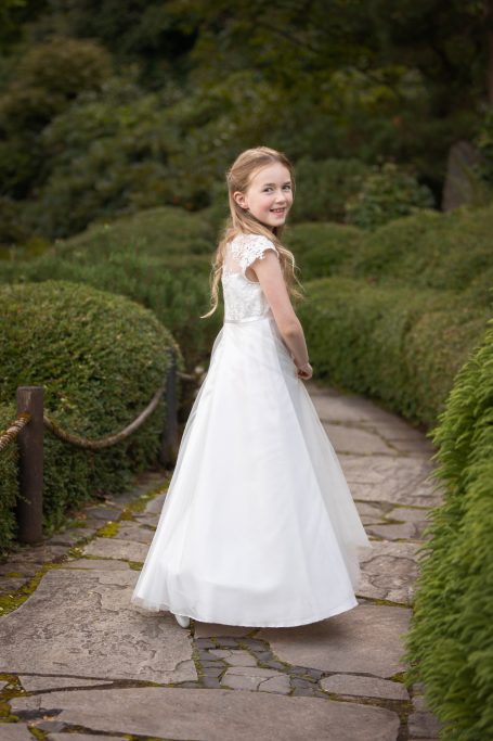 Ein Mädchen in einem weißen Kleid steht auf einem gepflasterten Weg in einer grünen Umgebung.