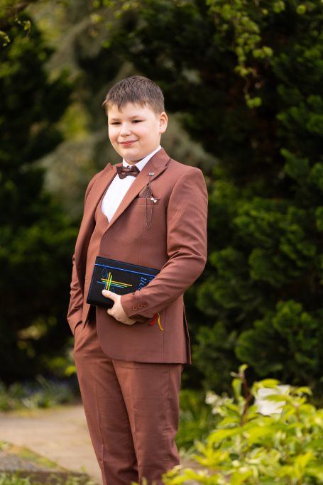 Junge in braunem Anzug mit Fliege hält ein Buch und steht vor grünen Bäumen.
