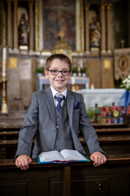 Ein Junge in Anzug steht an einer Holzbank in einer Kirche, lächelt und hält ein Buch.