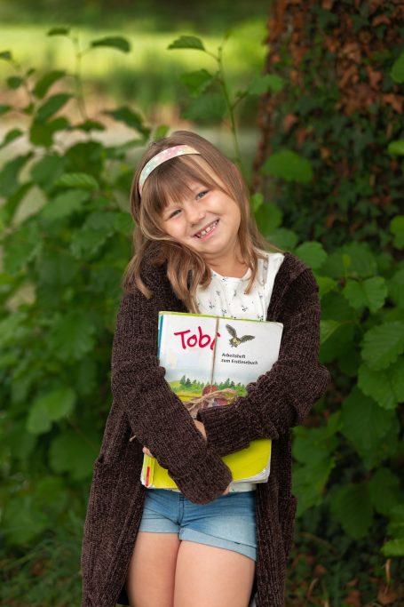 Lächelndes Mädchen mit einem Buch, umgeben von grünen Pflanzen.