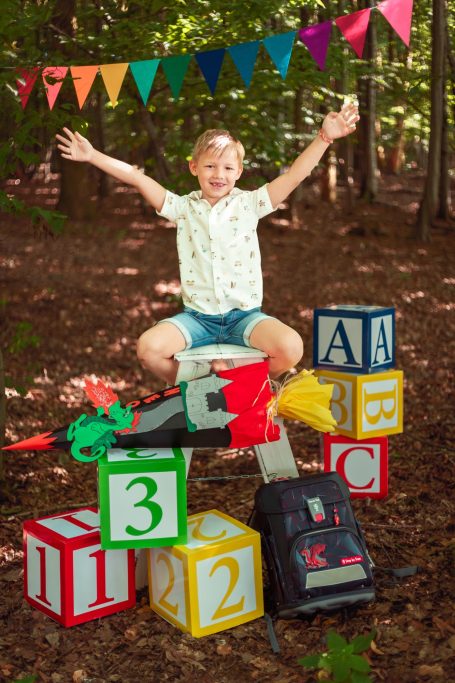 Ein Junge mit erhobenen Händen sitzt auf bunten Spielblöcken im Wald.