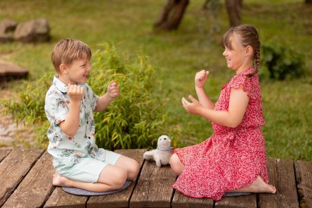 Zwei Kinder spielen auf einer Holzterrasse, umgeben von Grün, mit einem Spielzeug zwischen ihnen.