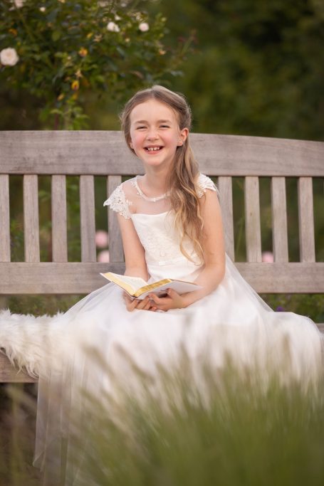 Ein Mädchen in einem weißen Kleid sitzt auf einer Bank und lächelt, während es ein Buch hält.