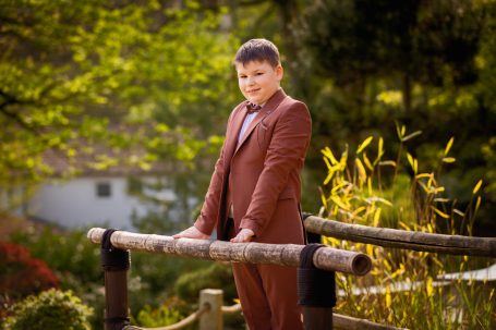 Junger Junge in einem braunen Anzug steht an einer Holzbrüstung in einer grünen Umgebung.