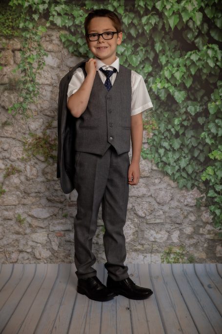 Ein kleiner Junge in Anzug mit Weste und Krawatte, lächelt und hält ein Jacket.