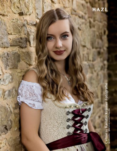 Junge Frau mit langen, lockigen Haaren, trägt ein traditionelles Kleid vor einer Steinmauer.