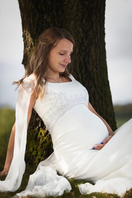 Schwangere Frau in einem weißen Kleid lehnt an einem Baum, mit sanfter Ausstrahlung.