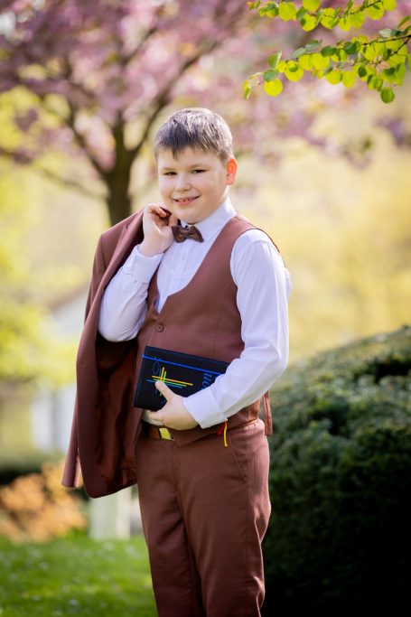 Ein Junge in einem braunen Anzug und Fliege hält eine Tasche und lächelt im Freien.
