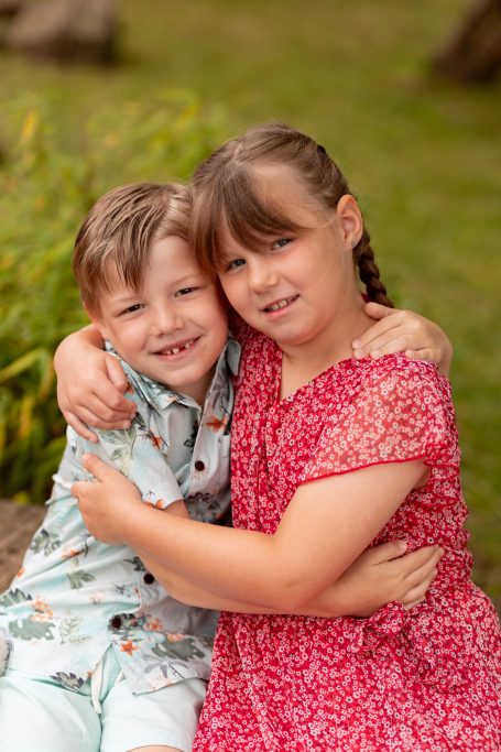 Ein Junge und ein Mädchen umarmen sich lächelnd im Freien, umgeben von Pflanzen.