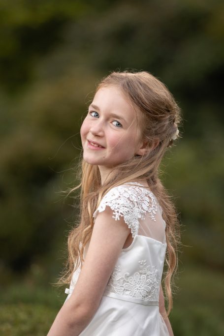 Mädchen mit langen, blonden Haaren und weißem Kleid, lächelt freundlich in der Natur.