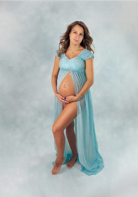 Schwangere Frau in einem blauen Kleid, umarmt ihren Bauch vor einem hellen Hintergrund.