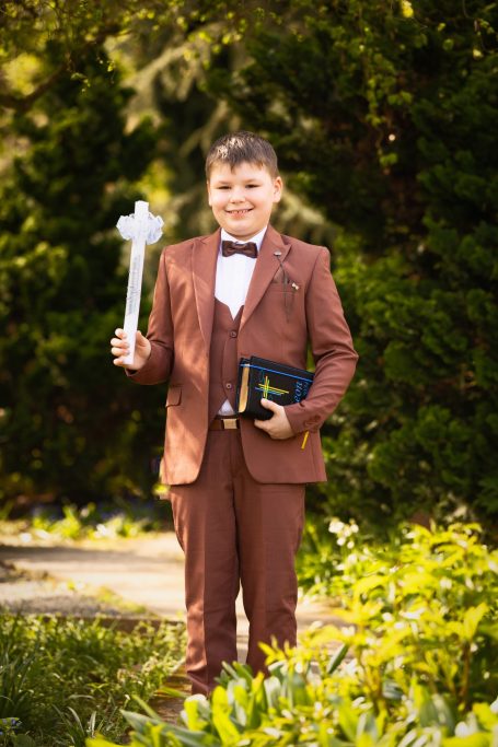 Ein Junge in einem braunen Anzug hält ein Kreuz und eine Bibel.
