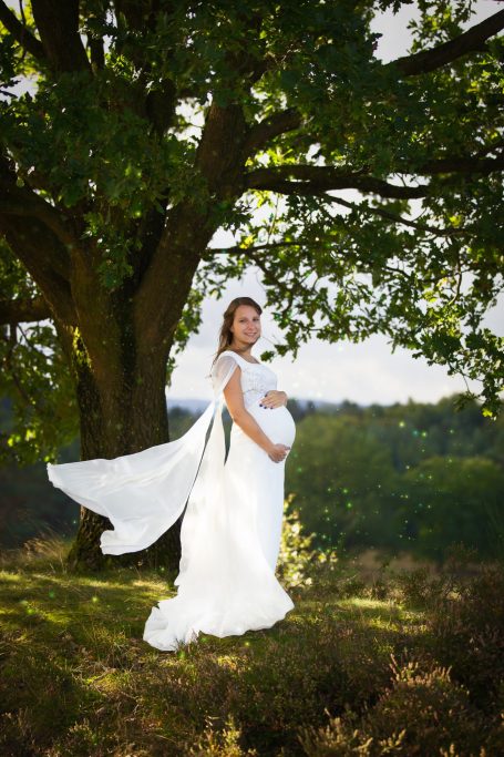Schwangere Frau in einem weißen Kleid steht unter einem großen Baum in der Natur.