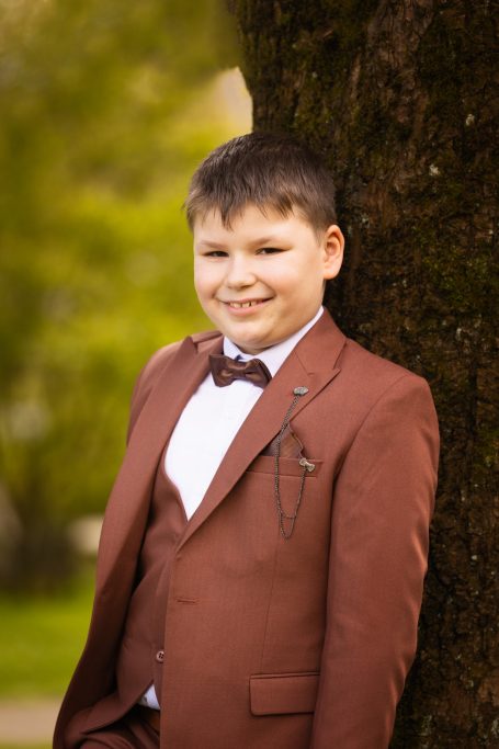 Ein lächelnder Junge in einem braunen Anzug steht an einem Baum.