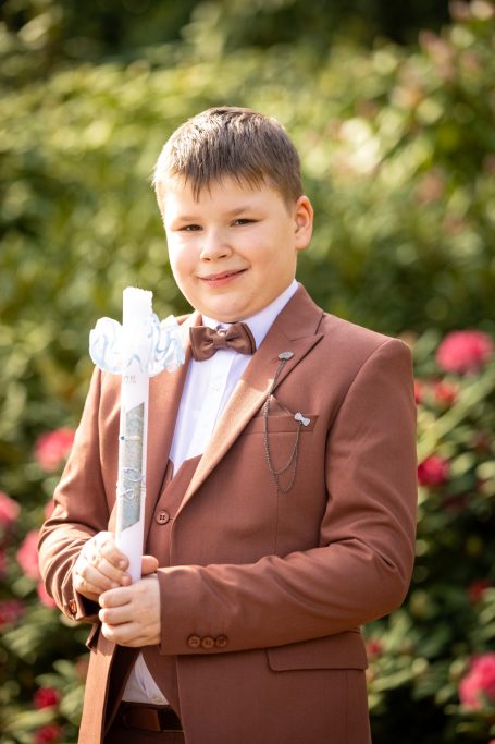 Ein Junge in einem braunen Anzug mit einer Fliege hält einen weißen Stock, umgeben von Blumen.