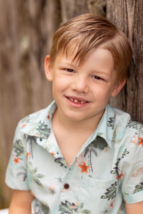 Ein lächelnder Junge mit blonden Haaren und floralem Hemd steht vor einer Holzlatte.