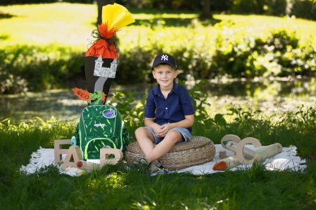 Junge sitzt im Freien neben einem bunten Rucksack und Spielzeug auf dem Rasen.