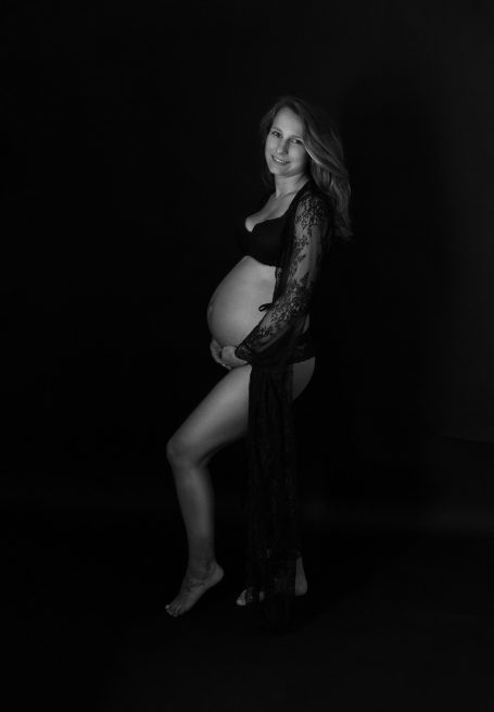 Schwangere Frau in eleganter Pose, umgeben von dunklem Hintergrund.