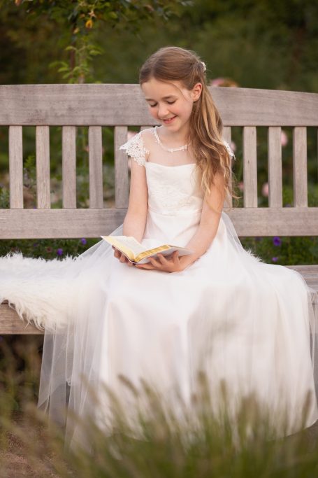 Ein Mädchen in einem weißen Kleid sitzt auf einer Bank und liest ein Buch.