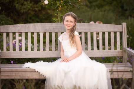 Ein Mädchen in einem weißen Kleid sitzt auf einer Bank in einem Garten.