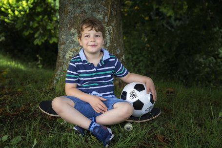Junge sitzt auf einem Skateboard, hält einen Fußball, umgeben von Bäumen und Gras.