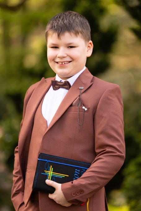 Ein Junge in Anzug mit Fliege hält eine Clutch und lächelt freundlich.