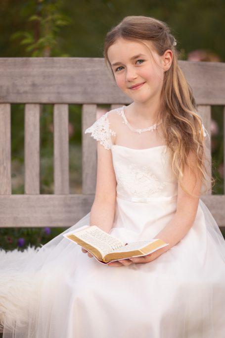 Mädchen in weißem Kleid sitzt auf einer Bank und hält ein aufgeschlagenes Buch.