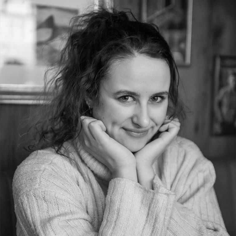 Eine lächelnde Frau mit lockigem Haar, die freundlich die Hände unter dem Kinn stützt.